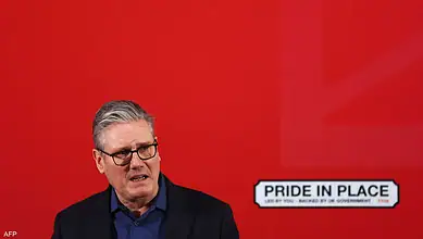 Politician speaking at a press conference with a "Pride in Place" sign in the background.