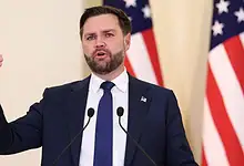 Politician delivering speech at a press event, emphasizing key policies with American flags behind h.