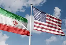 Iran and US flags flying side by side against a blue sky.